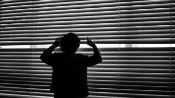 طفل يقف أمام نافذة مغلقة بستائر، يمد يديه نحو الأفق في إضاءة سوداء وبيضاء، يعبر عن الفضول والترقب.