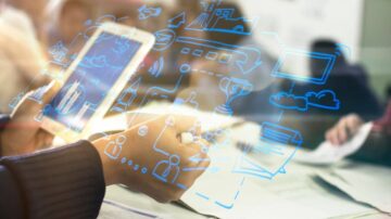 يد تظهر يدًا تمسك بجهاز لوحي مع رسومات رمزية زرقاء تمثل بيانات وابتكار، بينما يتم مناقشة مستندات على طاولة في الخلفية. تعكس الصورة تكنولوجيا المعلومات والتعاون في بيئة عمل حديثة.