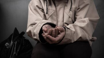 يدان فارغتان لشخص يرتدي سترة بيج، يجلس بجانب كيس قمامة في خلفية رمادية، مع تعبير يعكس الحزن أو الإحباط.