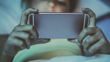 طفل في السرير يستخدم الهاتف المحمول ليلًا، مع التركيز على الشاشة في غرفة مظلمة.