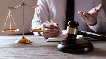 محامي يرتدي قميصًا أبيضًا يجلس على مكتب، أمامه ميزان العدالة ومطرقة قانونية. يقوم بتدوين ملاحظات في دفتر أثناء مناقشة موضوع قانوني. الصورة تعكس جوانب مهنية في مجال القانون.