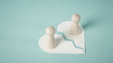 تمثالان خشبيان صغيران يقفان على طرفي قلب مقطوع، مع خلفية زرقاء فاتحة. يرمز المشهد إلى الانفصال أو العلاقات المتعطلة.