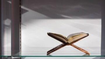 كتاب مفتوح على حامل خشبي، موضوع على رف زجاجي، مع ظلال خفيفة تسلط الضوء على التصميم البسيط.