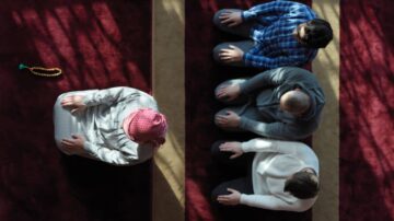 رجل يرتدي ملابس تقليدية يؤم مجموعة من الرجال خلال الصلاة في مسجد. يتواجدون على سجادة صلاة حمراء، مع تسليط الضوء على الأجواء الروحية والتركيز الجماعي على العبادة.