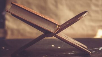 كتاب مفتوح مستند على حامل خشبي بتصميم بسيط، يظهر تفاصيل غلاف الكتاب بلون داكن. الخلفية غير واضحة، مما يبرز الكتاب والحامل.