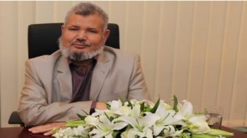 رجل في بدلة رمادية يجلس على مكتب خلفه ستارة بيضاء. لديه لحية رمادية ويبتسم. يوجد باقة من الزهور البيضاء أمامه وكأس ماء بجانبه.