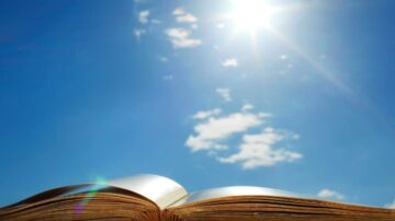 كتاب مفتوح تحت سماء زرقاء مشمسة مع غيوم بيضاء. الضوء ينعكس على صفحات الكتاب، مما يخلق تأثيراً ساحراً.