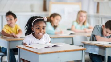 طفلة مبتسمة تجلس على مكتب في فصل دراسي، محاطة بأطفال آخرين يكتبون في دفاترهم. يوضح المشهد بيئة تعليمية ملهمة تعزز التعلم والتفاعل.