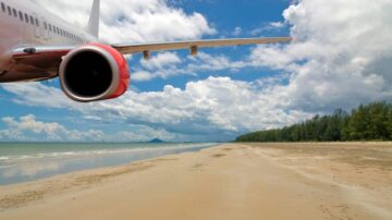 طائرة في السماء فوق شاطئ رملي مع مياه زرقاء وأشجار خضراء في الخلفية. السماء ملبدة بالغيوم مع أشعة الشمس.