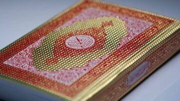 كتاب القرآن الكريم بتصميم ملون مع غلاف ذهبي وأحمر مزخرف. يبرز التصميم الفني الجميل للآيات القرآنية.