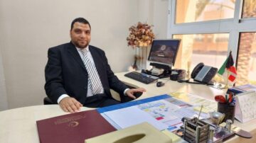 رجل في بدلة رسمية يجلس على مكتب في مكتب حديث. خلفه يوجد حاسوب شخصي وشاشة تعرض صورة، مع زهور صناعية وزينة مكتبية. يحتوي المكتب على مستندات وملفات متنوعة، مع العلمين الكويتي والعراقي ظاهرين.