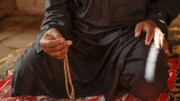 رجل يجلس على سجادة تقليدية ممسكًا بمسابح، يرتدي ملابس داكنة. الإضاءة تسلط الضوء على يده والمسابح، مما يبرز اللحظة الروحية.