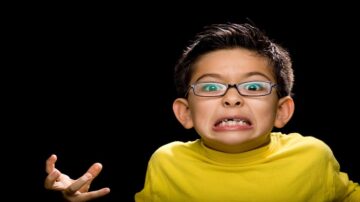 طفل يرتدي قميصًا أصفر، يظهر تعبير وجهه الغاضب والمرتبك، مع نظارات، وخلفية سوداء.