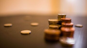 صورة لعملات معدنية متنوعة مرتبة بشكل غير منتظم على سطح خشبي، مع تركيز على أكوام العملات المختلفة. تشمل العملات عملات نحاسية وفضية بأحجام مختلفة.