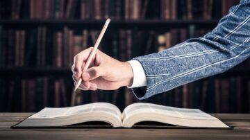 يدٌ تمسك قلم رصاص وتكتب على صفحة مفتوحة من كتاب، مع خلفية تحتوي على رفوف مليئة بالكتب، تعكس جوًا أكاديميًا.
