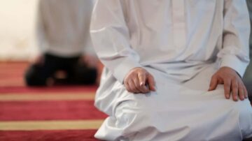 رجل يرتدي ثوباً أبيض يجلس على سجادة صلاة حمراء، بينما يظهر شخص آخر في الخلفية جالساً على ركبتيه. تعبر الصورة عن لحظة من الصلاة أو التأمل في بيئة دينية.