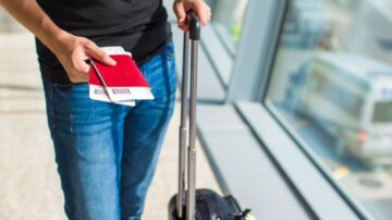 رجل يحمل حقيبة سفر صغيرة وجواز سفر وتذاكر طيران، يقف في مطار بالقرب من نافذة كبيرة.