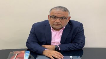 رجل في منتصف العمر يجلس على مكتب أمام جهاز كمبيوتر محمول، يرتدي بدلة داكنة وقميص وردي. يضع يده على الطاولة بجوار دفتر ملاحظات وقلم، ويظهر تعبير وجهه الجاد.