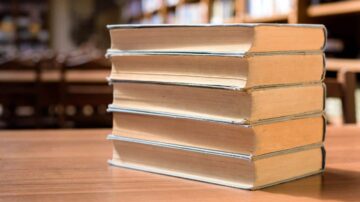 كتب مرتبة بشكل عمودي على طاولة خشبية في مكتبة، مع خلفية تحتوي على رفوف كتب.