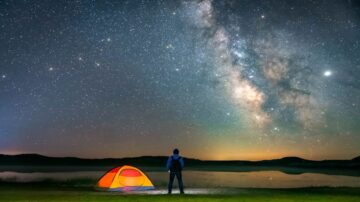 شخص يقف أمام خيمة ملونة تحت سماء مليئة بالنجوم، مع مجرة درب التبانة تضيء في الخلفية، يعكس جمال الطبيعة والليل في منطقة نائية.