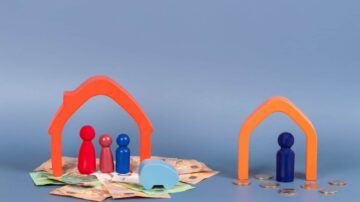مجسمات ملونة تمثل عائلة تحت سقفين برتقاليين، مع نقود ورقية وعملات معدنية في الخلفية. توضح الصورة مفهوم الادخار والاستثمار في المنازل.