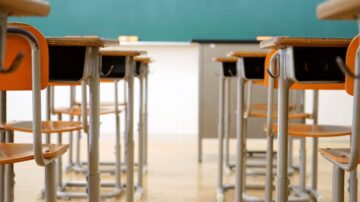 فصل دراسي مع مقاعد خشبية برتقالية وطاولة تعليمية، خلفية خضراء للسبورة. تصميم بسيط يبرز بيئة التعلم.