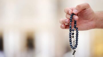 يد مرفوعة تحمل سبحة زرقاء داكنة مصنوعة من خرز لامع، مع تفاصيل فضية، في خلفية ضبابية توحي بمكان هادئ.