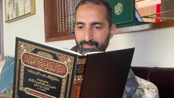 رجل يقرأ القرآن الكريم في غرفة معيشة مريحة، خلفه تظهر بعض الزخارف الإسلامية ونسخة أخرى من القرآن. تعكس الصورة أجواء من التركيز والتأمل الروحي.