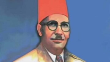 صورة لرجل يرتدي قبعة حمراء ونظارات، يظهر بابتسامة. الخلفية زرقاء، ويبدو أن الرجل يرتدي بدلة. تعكس الصورة طابعًا تاريخيًا وثقافيًا.