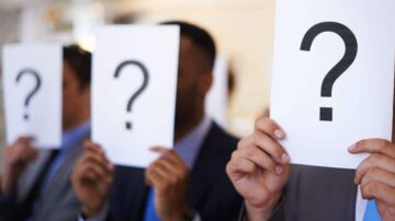 مجموعة من الأشخاص يرتدون بدلات رسمية، يحملون لوحات بيضاء بها علامة استفهام كبيرة، في بيئة داخلية. تعبر الصورة عن الغموض أو الاستفسار.