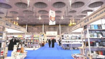 معرض للكتب يظهر أرفف مليئة بالكتب من مختلف الأنواع، مع ممرات واسعة وسقف مميز. الزوار يتجولون بين الأجنحة، حيث توجد لافتات توضح أرقام الأجنحة. الأرضية مغطاة بسجاد أزرق، مما يضيف لمسة جمالية للمكان.