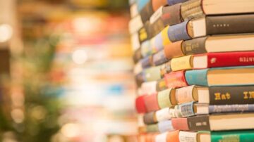 كتب ملونة مرتبة بشكل عمودي على رف، مع خلفية غير واضحة لمكتبة، تعكس أجواء القراءة والثقافة.