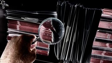 يد تظهر مكبرة تُفحص مجموعة من الملفات الورقية الملونة، مع التركيز على تفاصيل الأوراق والظلال. توضح الصورة عملية البحث والتدقيق في المعلومات.