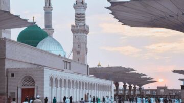 منظر خارجي لمسجد النبي في المدينة المنورة يظهر القبة الخضراء والمآذن. تقع المظلات الكبيرة في الساحة المحيطة، مع أشخاص يرتدون ملابس تقليدية. السماء تتلون بألوان الغروب.