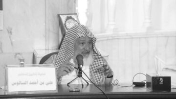 رجل مسن يرتدي غطاء رأس تقليدي يتحدث في مؤتمر أو ندوة. يظهر خلفه كتاب في إطار زجاجي. يركز على الحديث ويبدو جادًا.