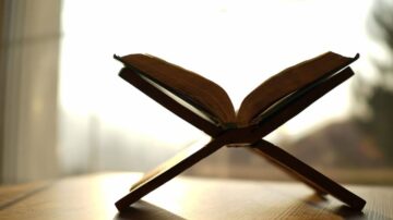 كتاب مفتوح موضوع على حامل خشبي، مع خلفية مضاءة. يظهر الكتاب بتفاصيل واضحة، مما يبرز صفحاته وأطرافه.