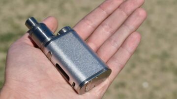 جهاز فاخر للتدخين الإلكتروني بحجم اليد، مصنوع من المعدن اللامع، مع تفاصيل دقيقة وفتحات تهوية. يظهر في يد شخص على خلفية طبيعية.