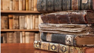 كتب قديمة مكدسة بشكل متراص على سطح خشبي، تظهر تفاصيل الغلاف والصفحات التالفة، مع خلفية تحتوي على رفوف كتب قديمة.