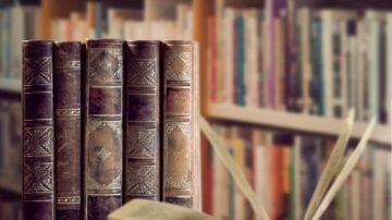 كتب قديمة مع غلاف جلدي، موضوعة أمام رفوف مكتبة تحتوي على مجموعة متنوعة من الكتب. التركيز على تفاصيل الغلاف والكتب المفتوحة في المقدمة.