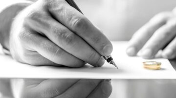 يد تظهر يدًا تكتب بقلم على ورقة بيضاء، مع وجود خاتم ذهبي بجوارها على سطح عاكس. تعكس الصورة لحظة من التوقيع أو الاتفاق، مما يوحي بمسائل قانونية أو شخصية.