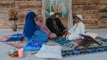 أسرة مسلمة تجلس معًا على سجادة ملونة في غرفة مزينة بأجواء دافئة. الأطفال يرتدون ملابس تقليدية، بينما الكبار يتبادلون الحديث حول القرآن. الإضاءة الطبيعية تضيء المكان، مع عناصر زخرفية مثل الوسائد والأقمشة.