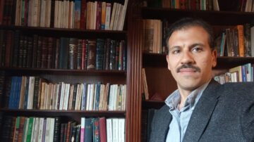 رجل يقف أمام مكتبة مليئة بالكتب، يرتدي سترة رمادية وقميص أزرق، يظهر تعبيره الواثق في إطار الصورة.