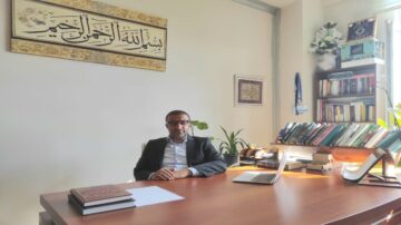 رجل يجلس على مكتب في مكتب أنيق، محاط بكتب دينية ونباتات. في الخلفية، لوحة مزخرفة تحمل آية قرآنية. يوجد لابتوب على المكتب وأوراق مرتبة.