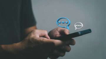 يد تحمل هاتفًا ذكيًا مع رموز فقاعات محادثة زرقاء وبيضاء، تعكس التواصل الرقمي والمحادثات النصية.