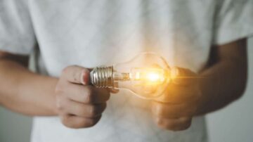 شخص يحمل مصباح كهربائي مضاء في يديه، مع توهج واضح ينبعث منه، مما يرمز إلى الأفكار المشرقة والإلهام.