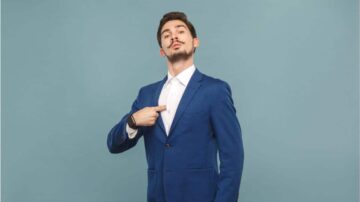 رجل يرتدي بدلة زرقاء وقميص أبيض، يشير بإصبعه إلى جيب بدلته، مع تعبير واثق، خلفية رمادية.