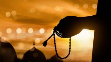 يد مرفوعة تحمل سبحة، silhouetted ضد غروب الشمس، مع خلفية مضببة. تعكس الصورة روحانية وهدوء.