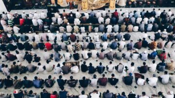 مجموعة من المصلين في مسجد، يجلسون على الأرض في صفوف مرتبة، يرتدون ملابس تقليدية. يظهر المسجد بتفاصيل معمارية جميلة، مع زخارف وألوان دافئة. الصلاة تعبر عن الوحدة والتركيز الروحي.