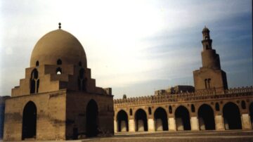 صورة لمجمع معماري تاريخي يضم قبة كبيرة ومنارة. القبة ذات شكل دائري وتتميز بلونها الرملي، بينما المنارة ترتفع بشكل أنيق. يُظهر المشهد سماء زرقاء فاتحة مع سحب خفيفة، مما يبرز التفاصيل المعمارية الجميلة للمكان.