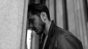 رجل يضع رأسه على جدار، يبدو عليه الإحباط والتفكير العميق، يرتدي سترة جلدية، مع خلفية معمارية ضبابية.